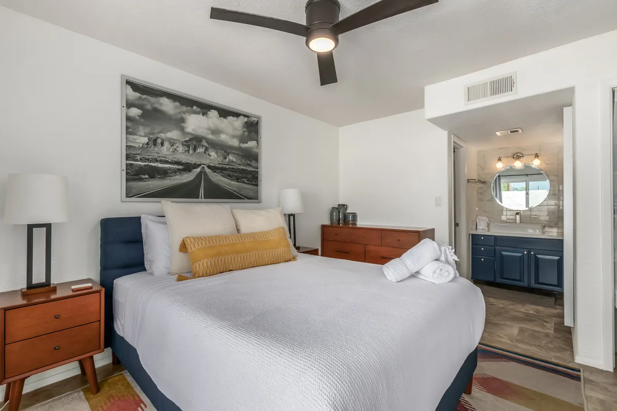 This image features a beautifully made bed with decorative pillows, framed landscape artwork on the wall, nightstands with soft-glow lamps, and a vanity area nearby. The warm color palette, soft lighting, and styled linens evoke a sense of both comfort and elegance, while the bedroom’s layout feels airy and welcoming.
