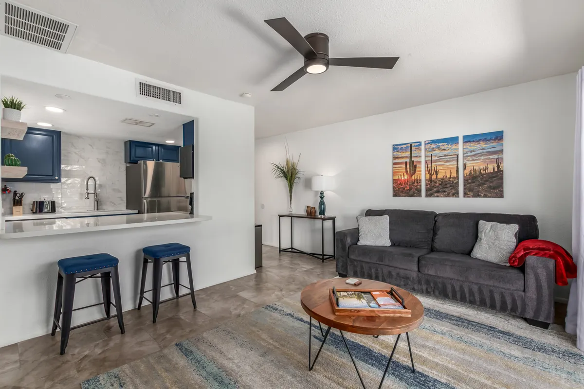 This image features a sweeping view of the living area and kitchen, adorned with striking blue cabinetry, a modern light fixture, desert-inspired artwork, and a coffee table with magazines. The plush designer sofa is prominently visible, complemented by contemporary décor and abundant natural light that gives the space a cozy yet vibrant feel.