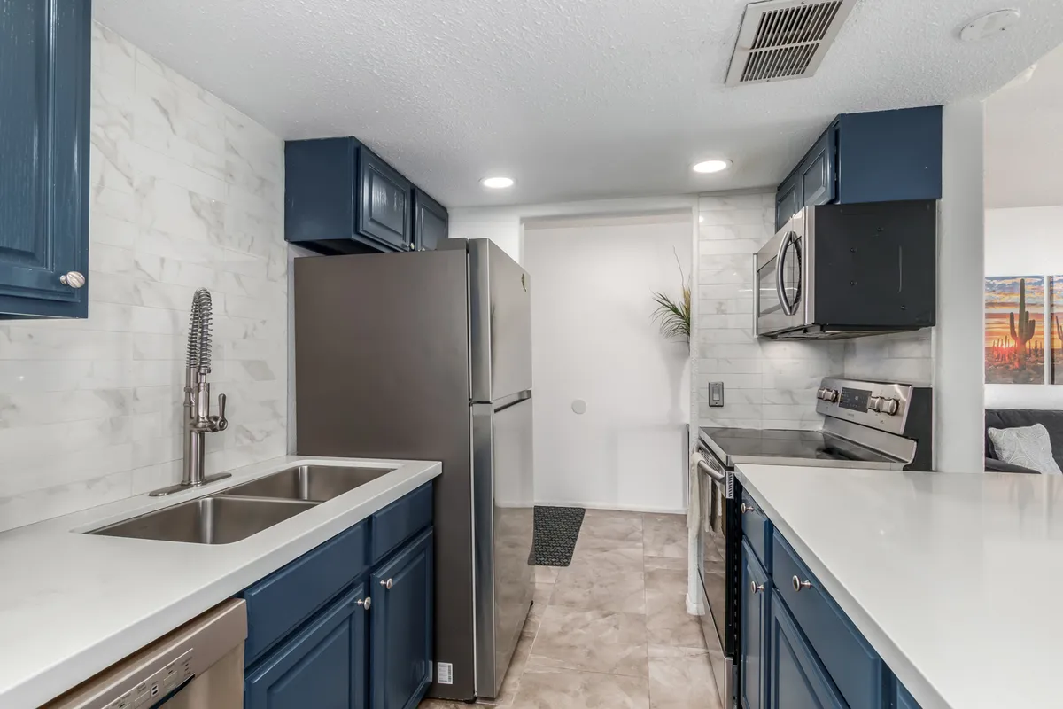 A warm, functional kitchen with blue cabinetry, stainless steel appliances, white countertops, and a textured backsplash. The scene feels both inviting and practical, suited for family meal preparation or social gatherings.