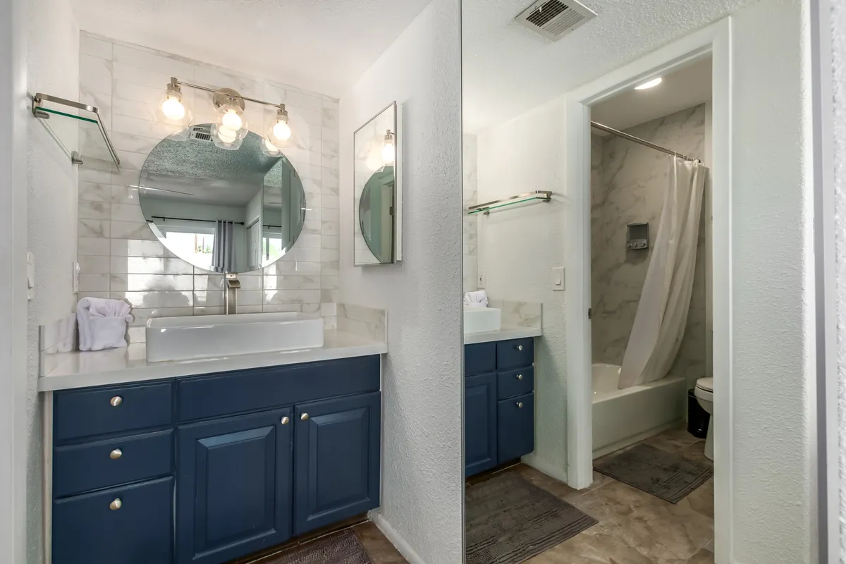 A stylish ensuite bathroom featuring a blue vanity, large round mirror, elegant light fixtures, and a pristine white bathtub. The space is well-lit and modern, with details that signal high-quality amenities and a spa-like experience.