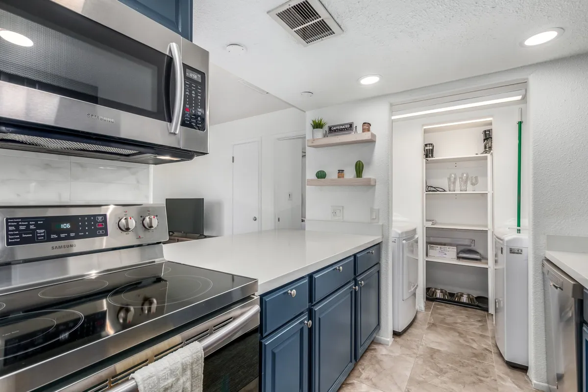 A focused view of the kitchen highlights gleaming stainless steel appliances, deep blue cabinetry, and crisp white countertops. The space looks both functional and modern, ideal for preparing meals or enjoying morning coffee rituals as the description suggests.