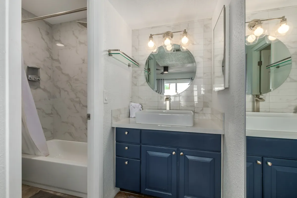 A clean, fresh bathroom features a round mirror above the sink, blue cabinetry, and elegant marble-like tiles. The atmosphere is serene and well-appointed, perfect for relaxing routines and easy settling in.